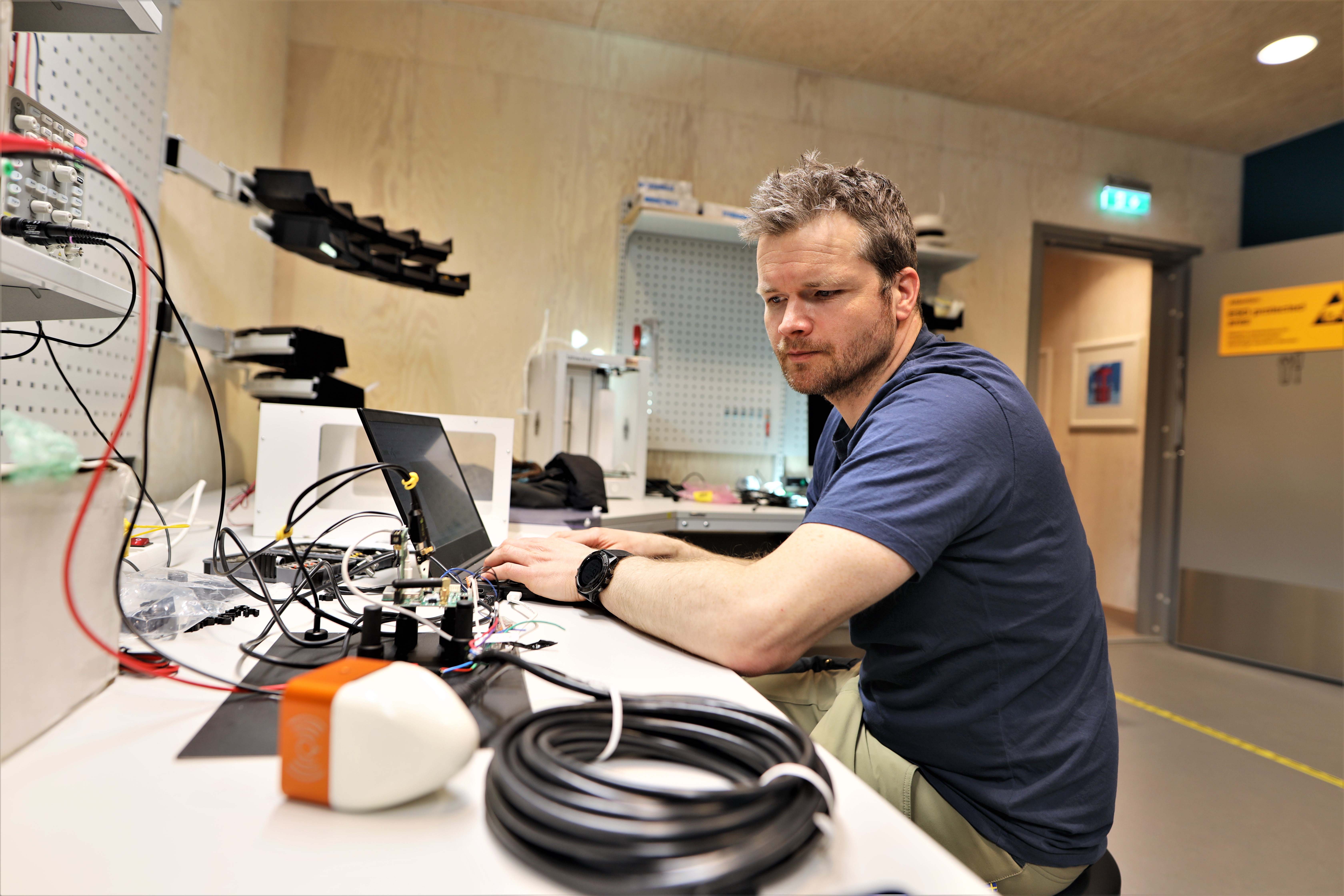 Man sitting in a tech lab looking at a IoT sensor while typing on laptop computer.